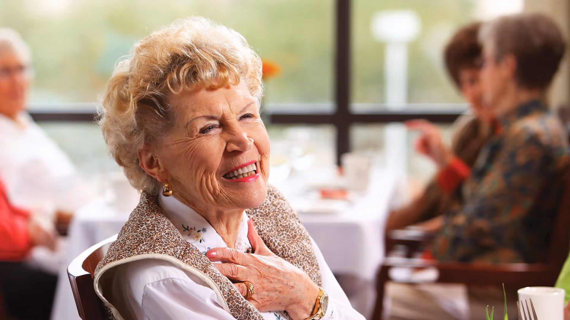 An elderly woman smiling and touching her chest while seated at a dining table with other seniors blurred in the background.