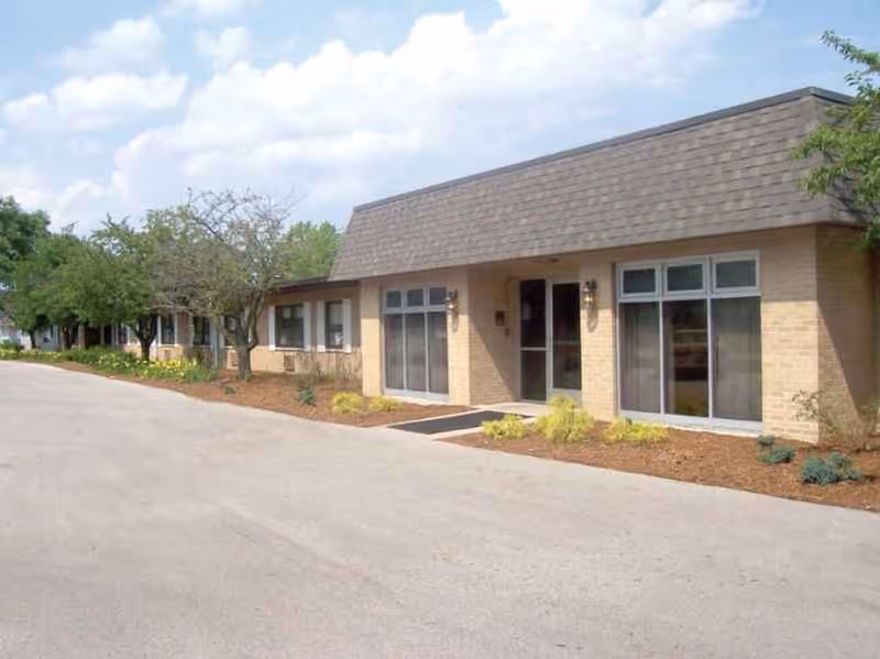 Exterior view of a single-story brick building with large windows and a shingled roof, surrounded by a paved driveway and landscaped with small bushes and trees under a partly cloudy sky.