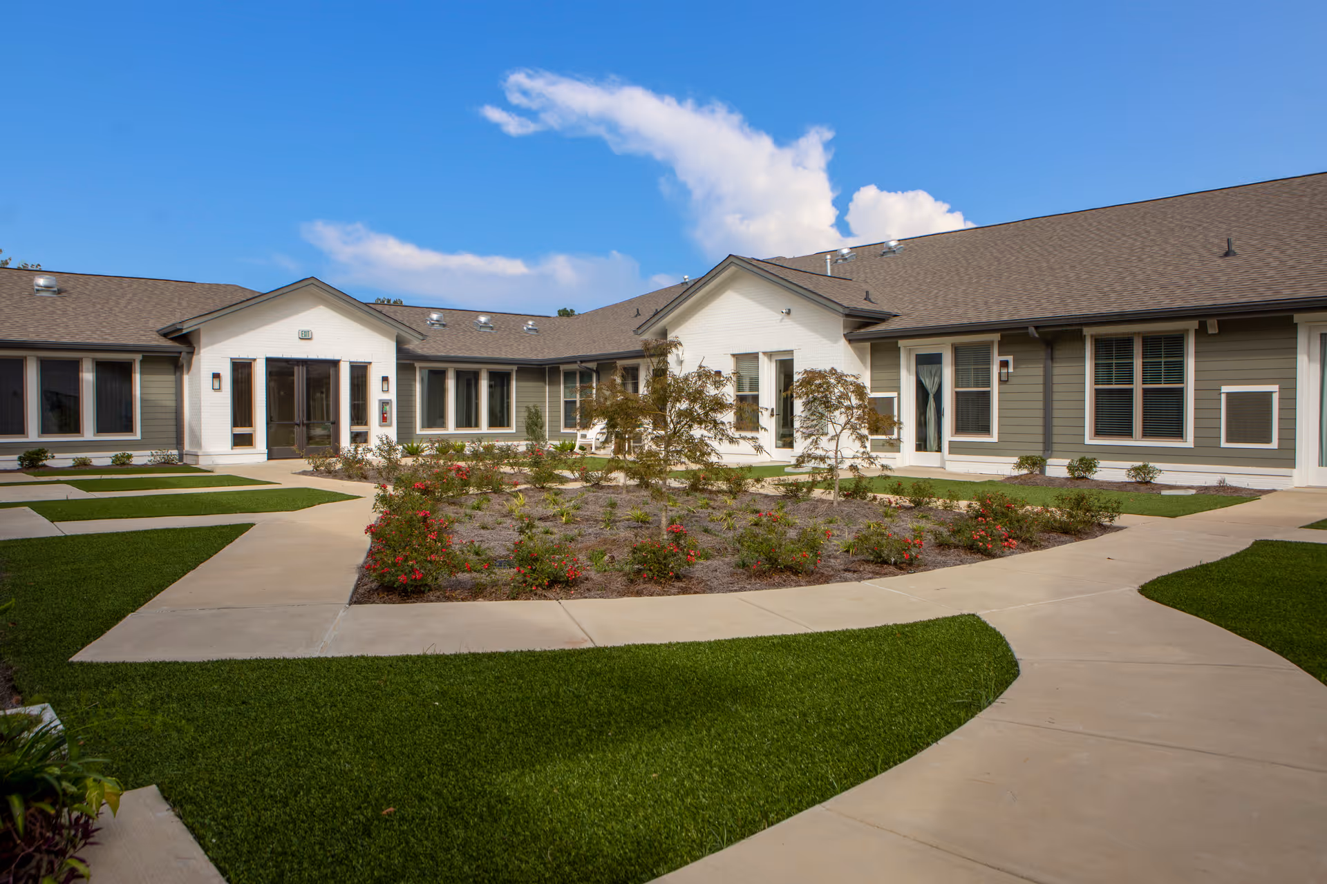 Exterior view of Avanti Senior Living at Lafayette showing a single-story building with a courtyard garden in the center. The courtyard has a paved walkway surrounding a landscaped area with small trees and flowering plants. The building has multiple windows and doors, with a clear blue sky in the background.