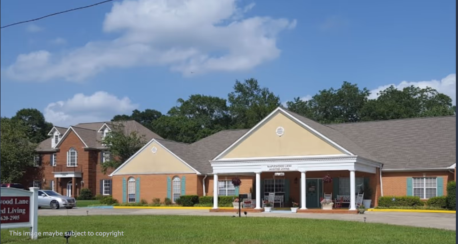 Front exterior view of Maplewood Lane Assisted Living facility, showing a single-story building with a covered entrance supported by white columns, surrounded by green grass and trees under a blue sky with some clouds.