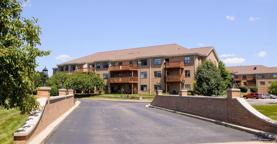 Exterior view of a three-story brick senior living facility with balconies, surrounded by trees and a paved driveway with brick walls on either side under a clear blue sky.