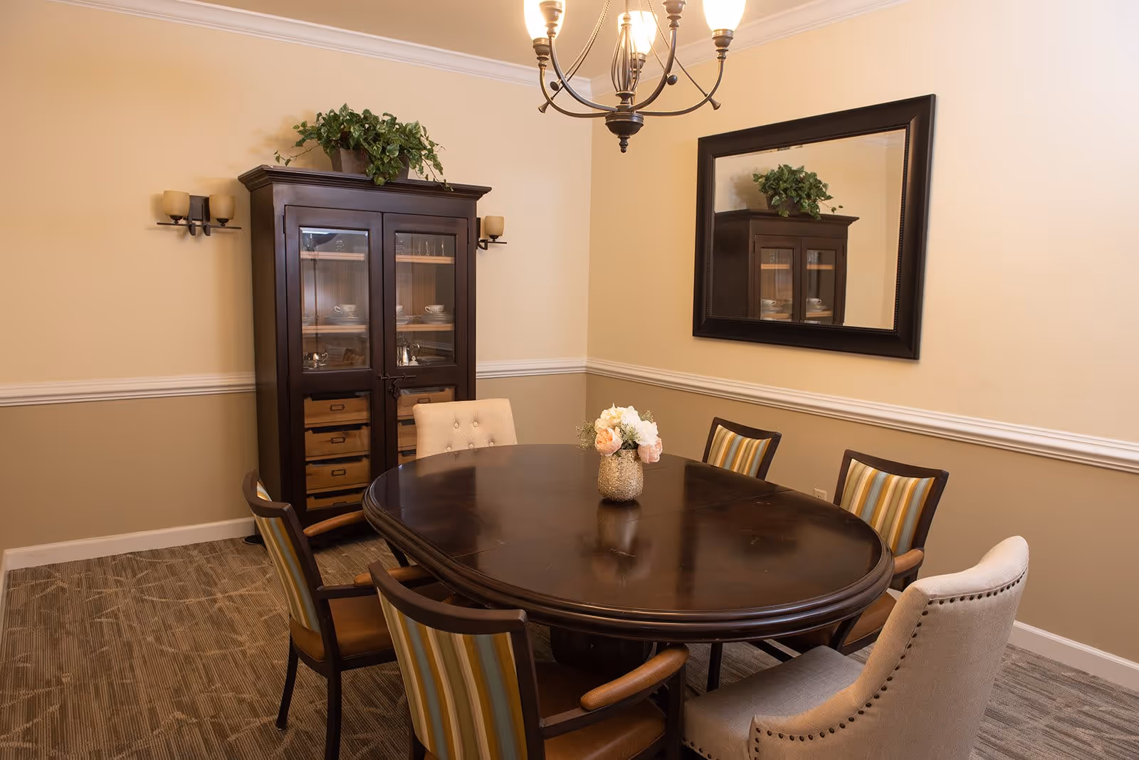 A dining room with a dark wooden oval table surrounded by six chairs, including two beige upholstered chairs and four wooden chairs with striped cushions. A vase with flowers is placed in the center of the table. Behind the table is a dark wooden cabinet with glass doors displaying dishes, topped with a green plant. A large rectangular mirror with a dark frame hangs on the wall, reflecting the cabinet. The walls are painted beige with white trim, and a chandelier with multiple lights hangs from the ceiling.