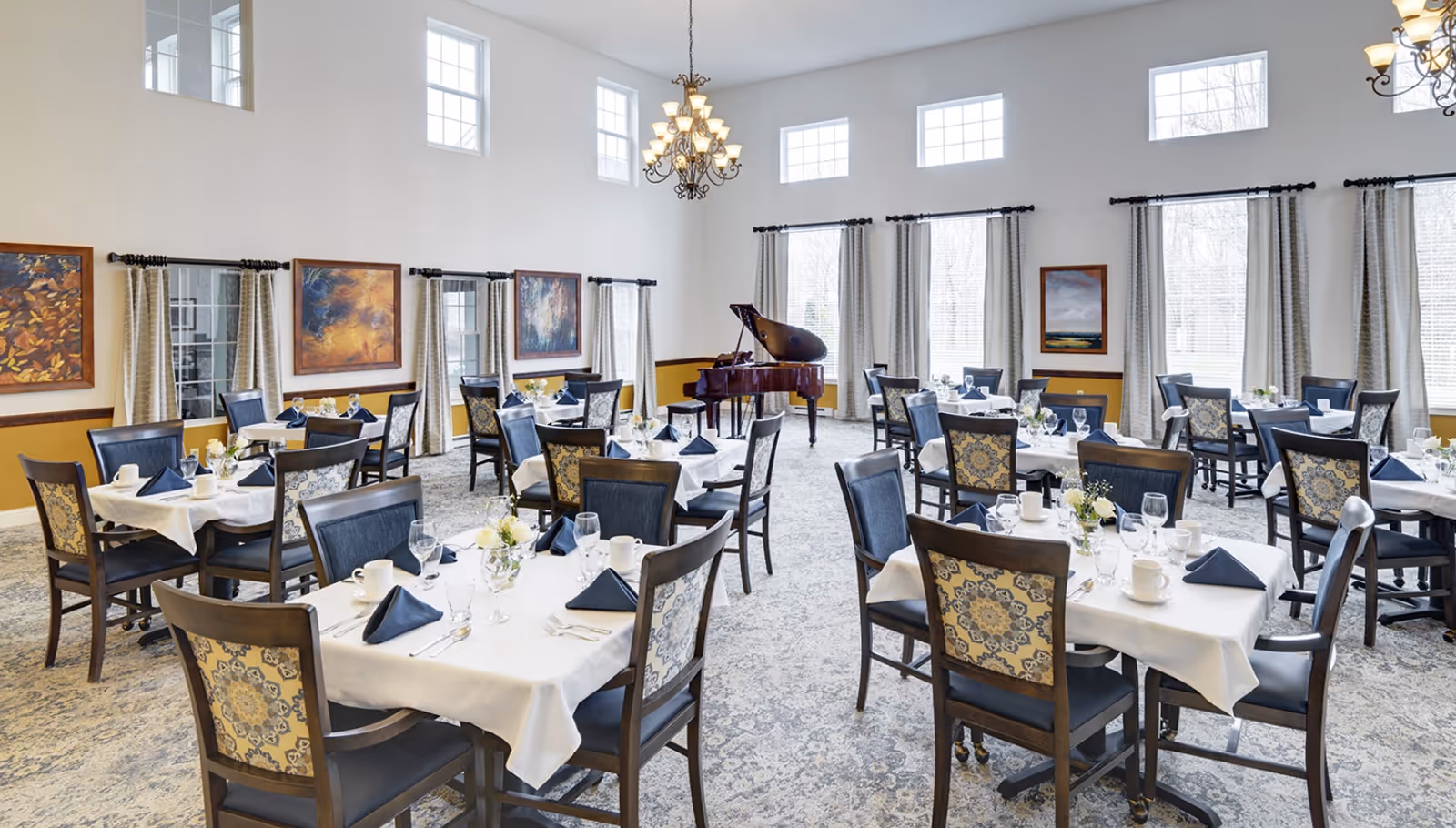 A bright and elegant dining room with multiple tables set with white tablecloths, navy blue napkins, glassware, and white cups. The room features large windows with curtains, several framed paintings on the walls, a grand piano in the corner, and a chandelier hanging from the ceiling.