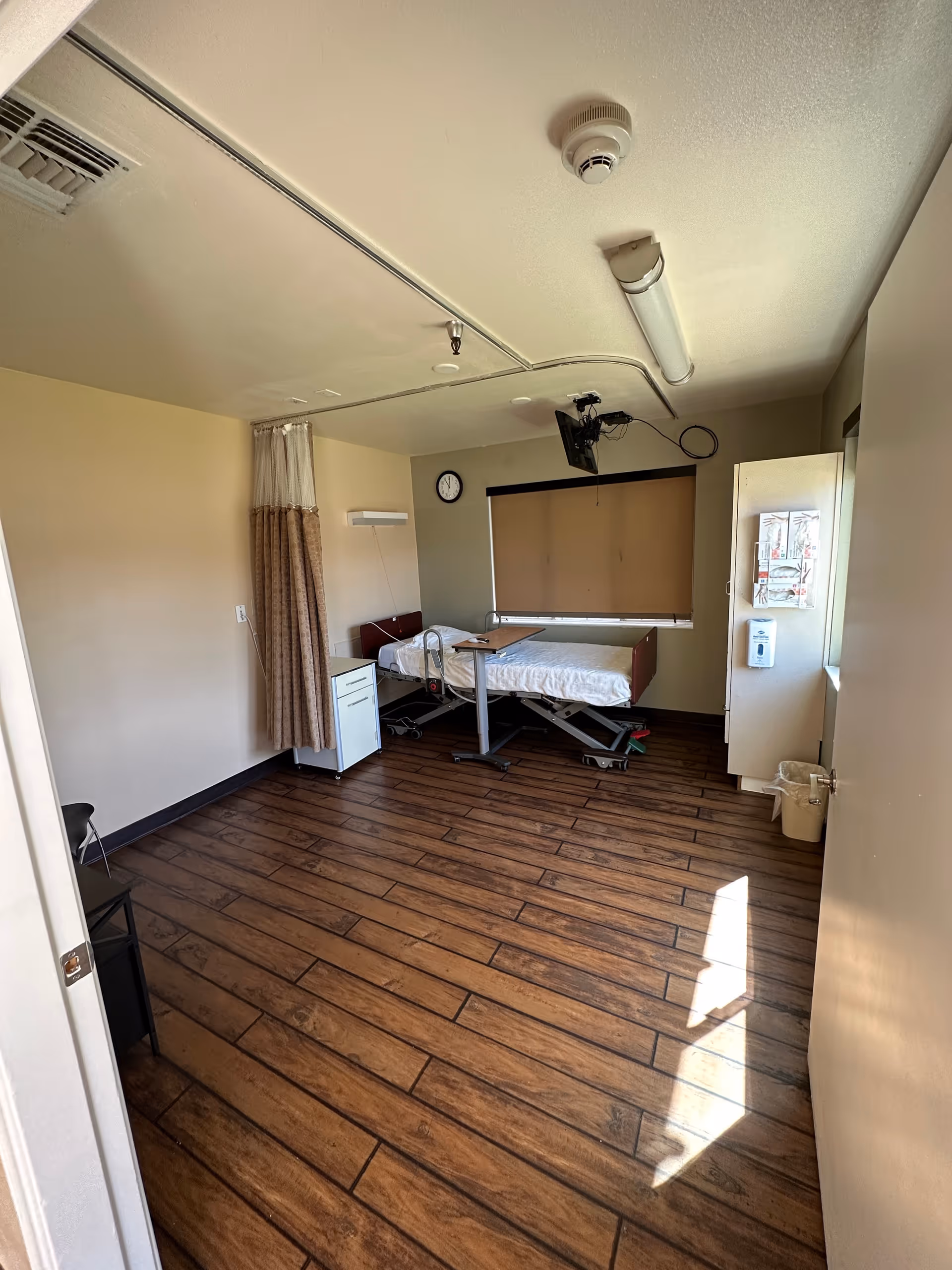 Empty nursing facility patient room with a single hospital bed, bedside cabinet, privacy curtain, wall clock and wood-look floor.