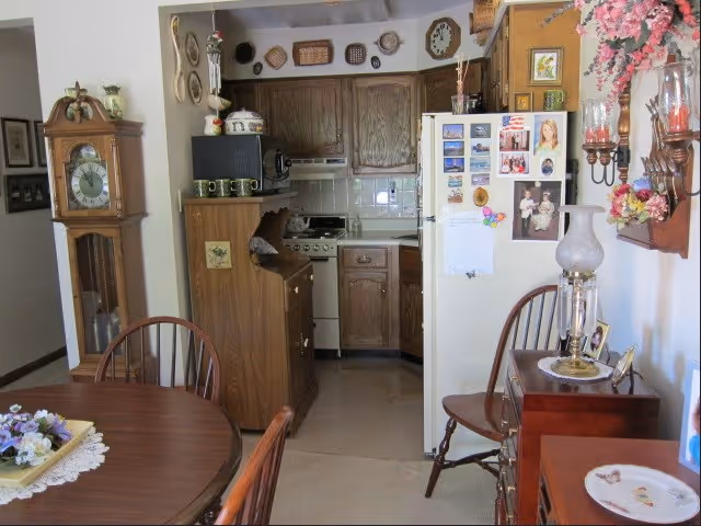 Dining area with a wooden table and chairs next to a small kitchen featuring oak cabinets, a white refrigerator covered in magnets, and a grandfather clock.