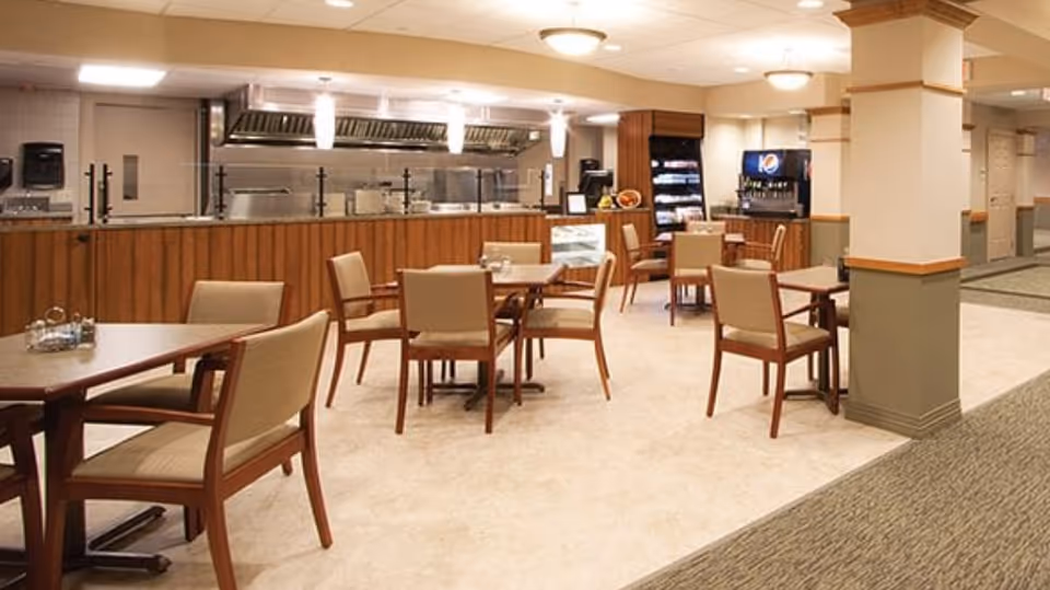 A dining area with several wooden tables and cushioned chairs arranged neatly. In the background, there is a serving counter with a glass sneeze guard, kitchen equipment, and a beverage dispenser. The room has beige walls, carpeted and tiled flooring, and ceiling lights.