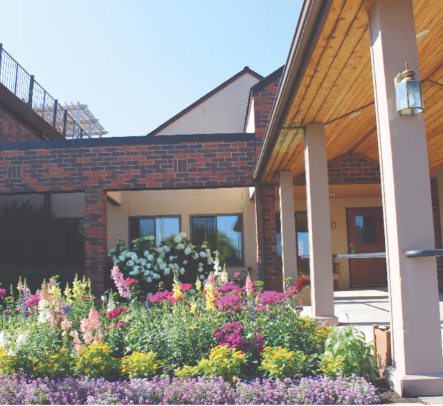 Entrance of a brick senior living building with a covered porch and colorful flower beds in front.