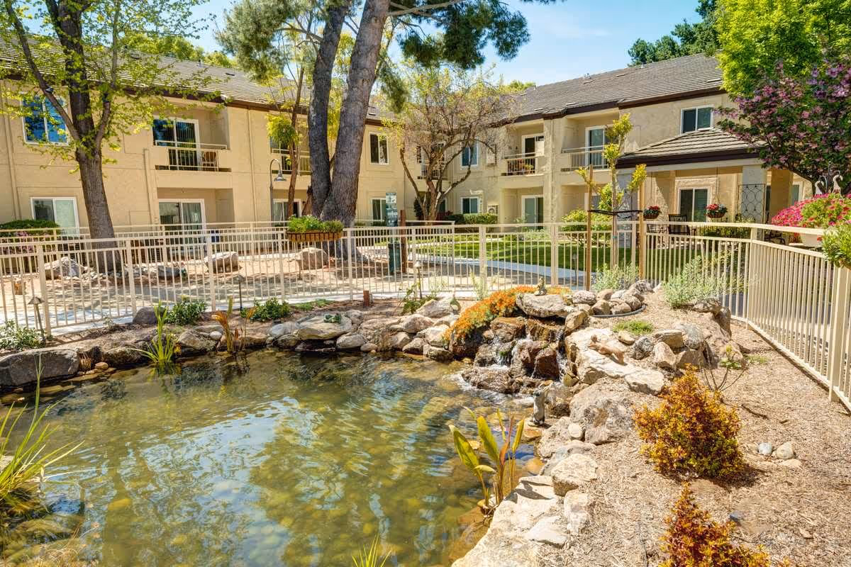 Outdoor view of The Oaks at Inglewood senior living facility showing a landscaped garden area with a small pond and waterfall surrounded by rocks and plants. The two-story building with balconies and windows is visible in the background under a clear blue sky.
