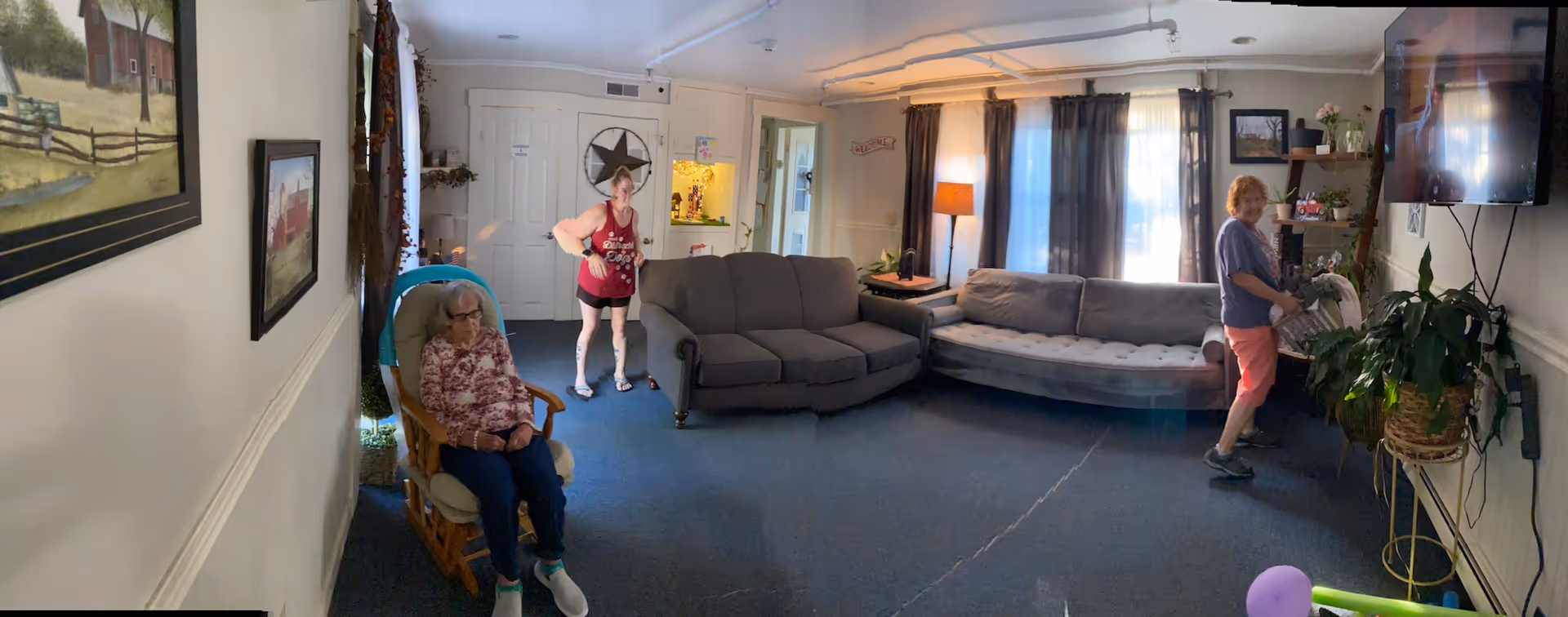 A cozy living room with two gray sofas, a rocking chair occupied by an elderly woman, and two other women standing and interacting. The room has blue carpet, a floor lamp, a wall-mounted TV, several plants, and framed pictures on the walls. Natural light comes through windows with dark curtains.