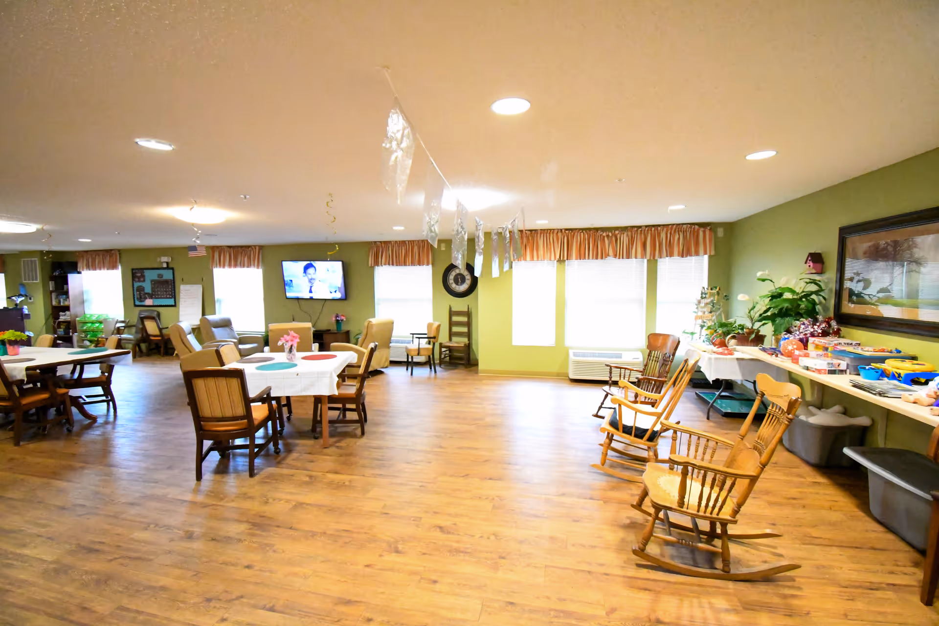 A spacious common area in a senior living facility with wooden flooring, several tables and chairs, rocking chairs along the right wall, a television mounted on the far wall, and windows with curtains letting in natural light. There are decorations hanging from the ceiling and a table with plants and games on the right side.