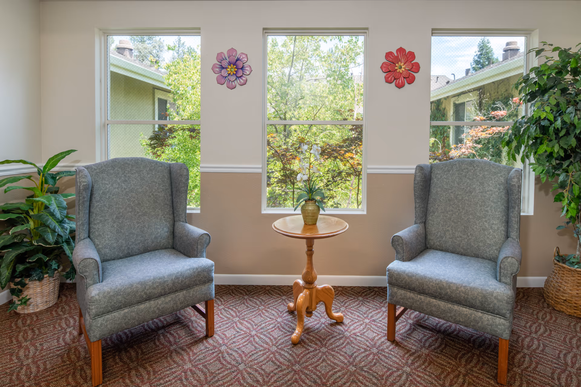 Two upholstered armchairs flank a small wooden table with a potted plant in front of three windows in a bright sitting area.