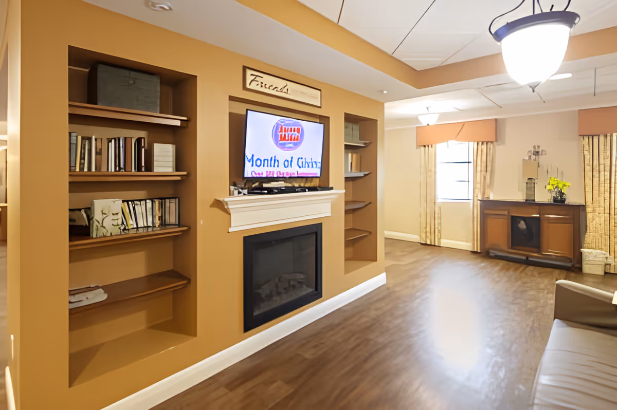 A cozy living room area with a built-in wall unit featuring shelves filled with books and decorative items. A flat-screen TV is mounted above a white mantel fireplace. The room has warm beige walls, wood flooring, and a ceiling light fixture. In the background, there is a window with curtains and a wooden cabinet with decorative items on top.