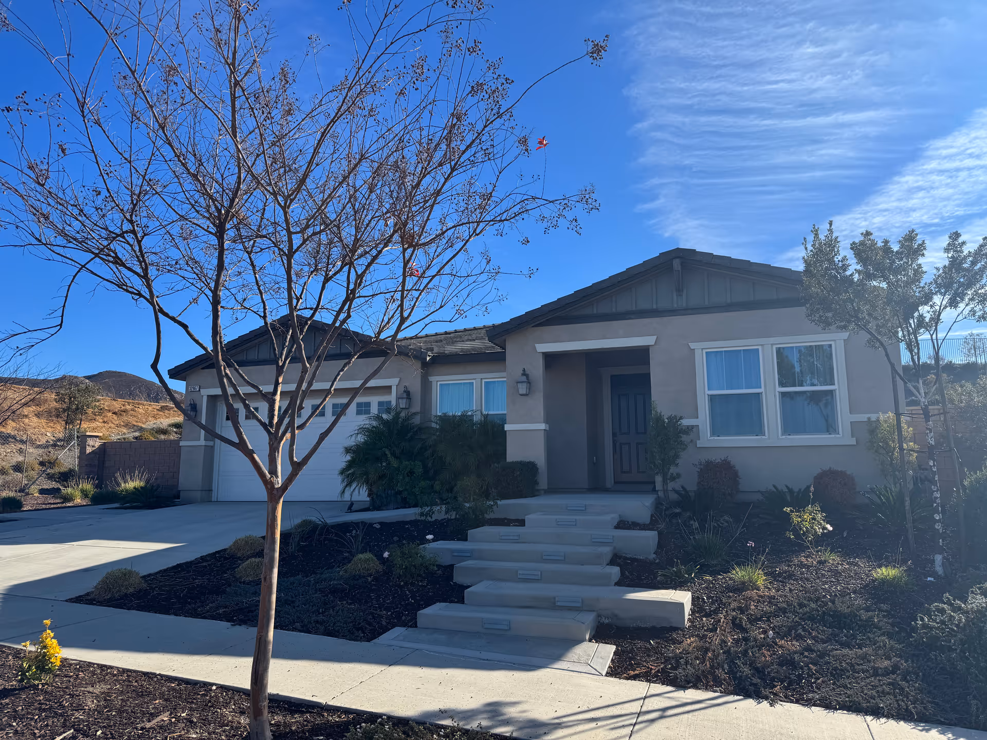 Exterior view of a single-story house with a front porch, steps leading up to the entrance, a garage, and landscaped yard with trees and shrubs under a clear blue sky.