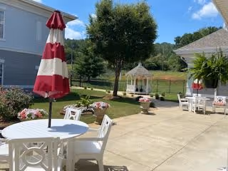 Outdoor patio area with white plastic tables and chairs, one table with a red and white striped umbrella. There are flower pots with pink and white flowers, a white gazebo in the background, green grass, trees, and a clear blue sky.