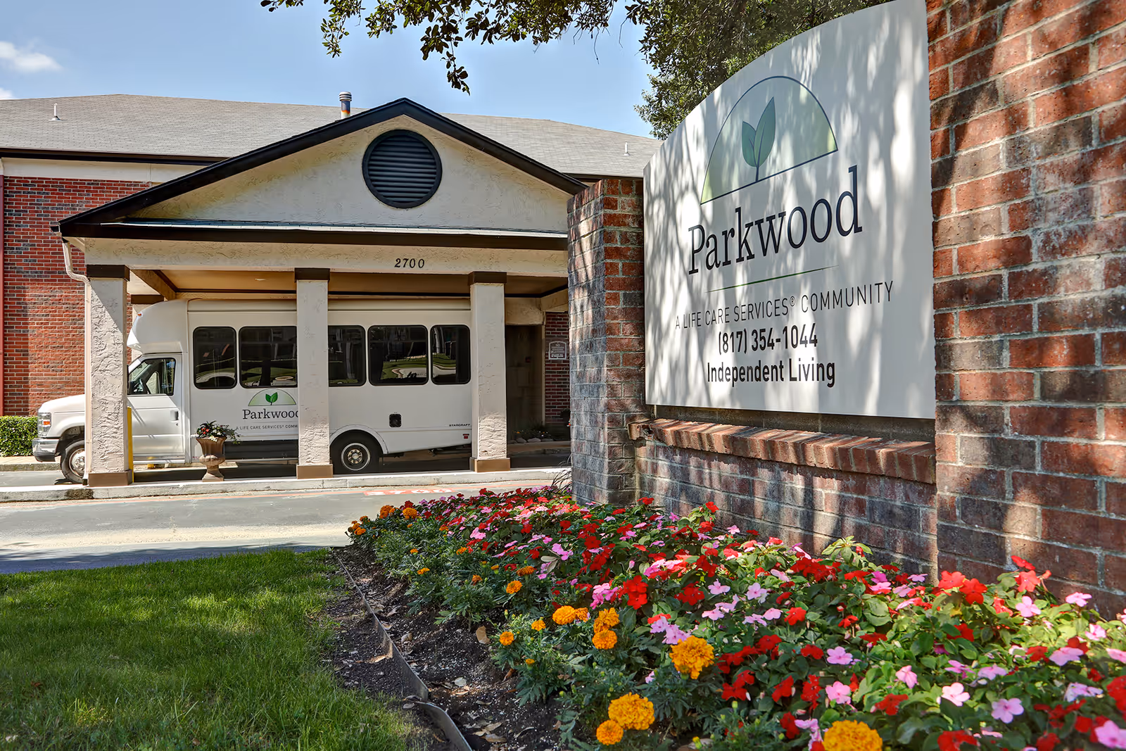 Exterior view of Parkwood Independent Living entrance with a shuttle bus and colorful flower beds.