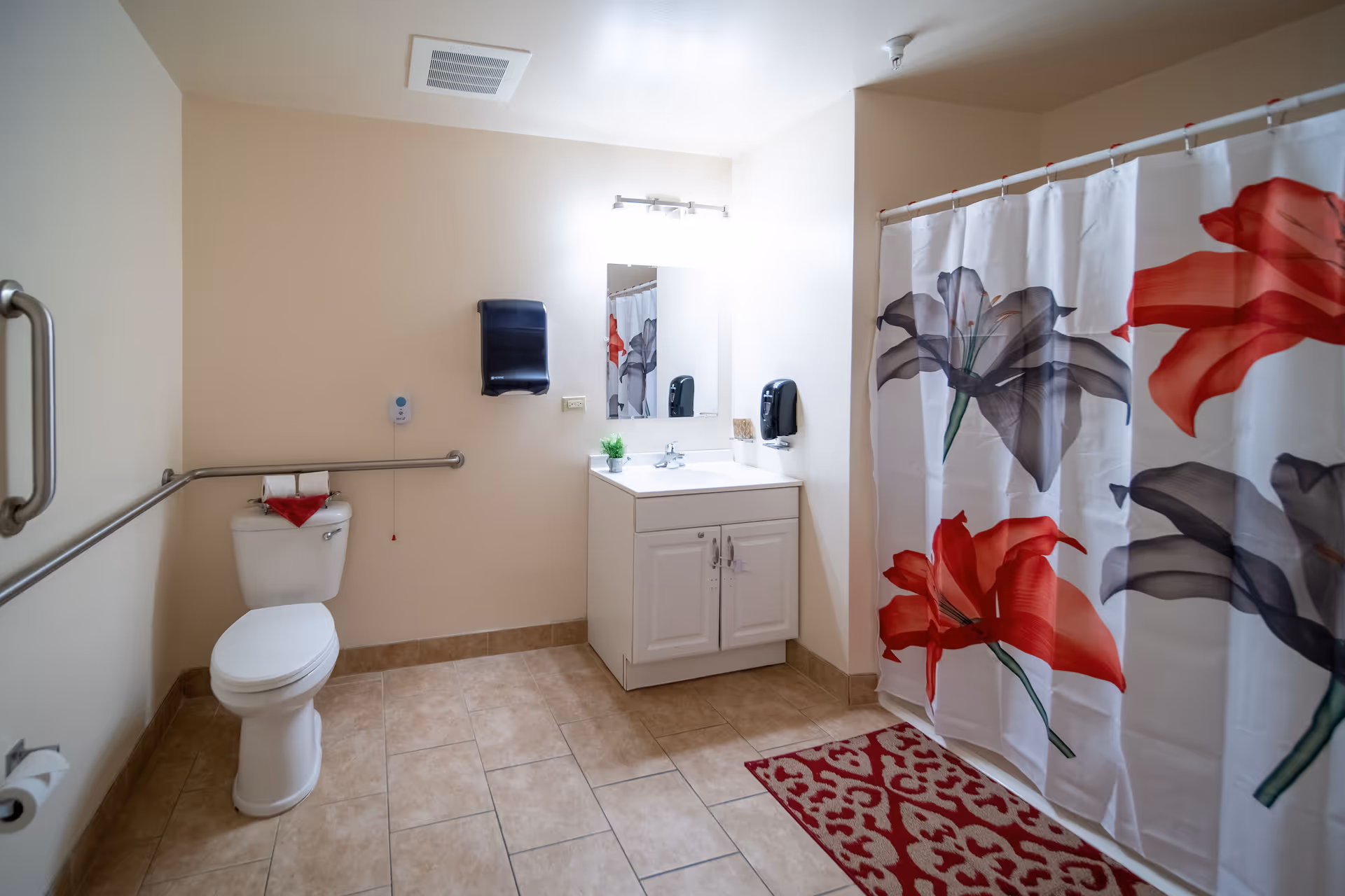 A clean and spacious bathroom with beige tiled floor and walls. There is a white toilet with a grab bar on the wall beside it, a white vanity with a sink and mirror above it, and a shower with a curtain featuring large red and gray floral designs. A red patterned rug is placed in front of the shower.