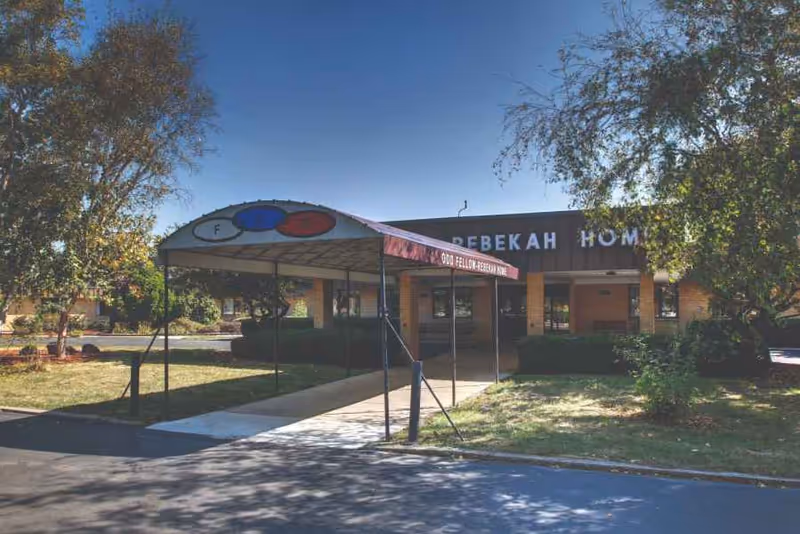 Front exterior view of the Odd Fellow-Rebekah Home building with a covered entrance canopy. The building is surrounded by trees and greenery under a clear blue sky.