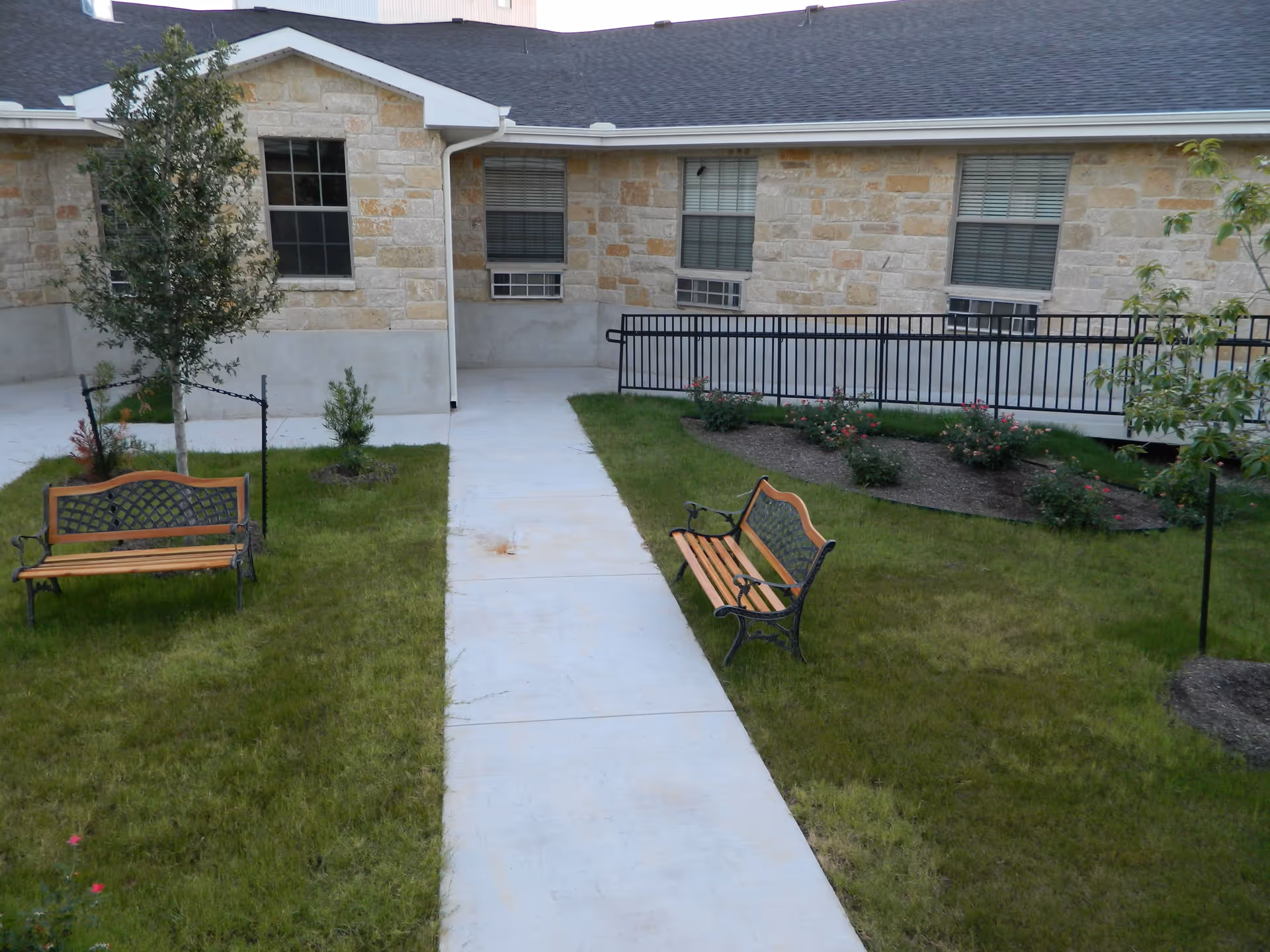 Outdoor courtyard area with a concrete walkway, two wooden benches with metal frames, green grass, small trees, and bushes in front of a stone building with windows and air conditioning units.