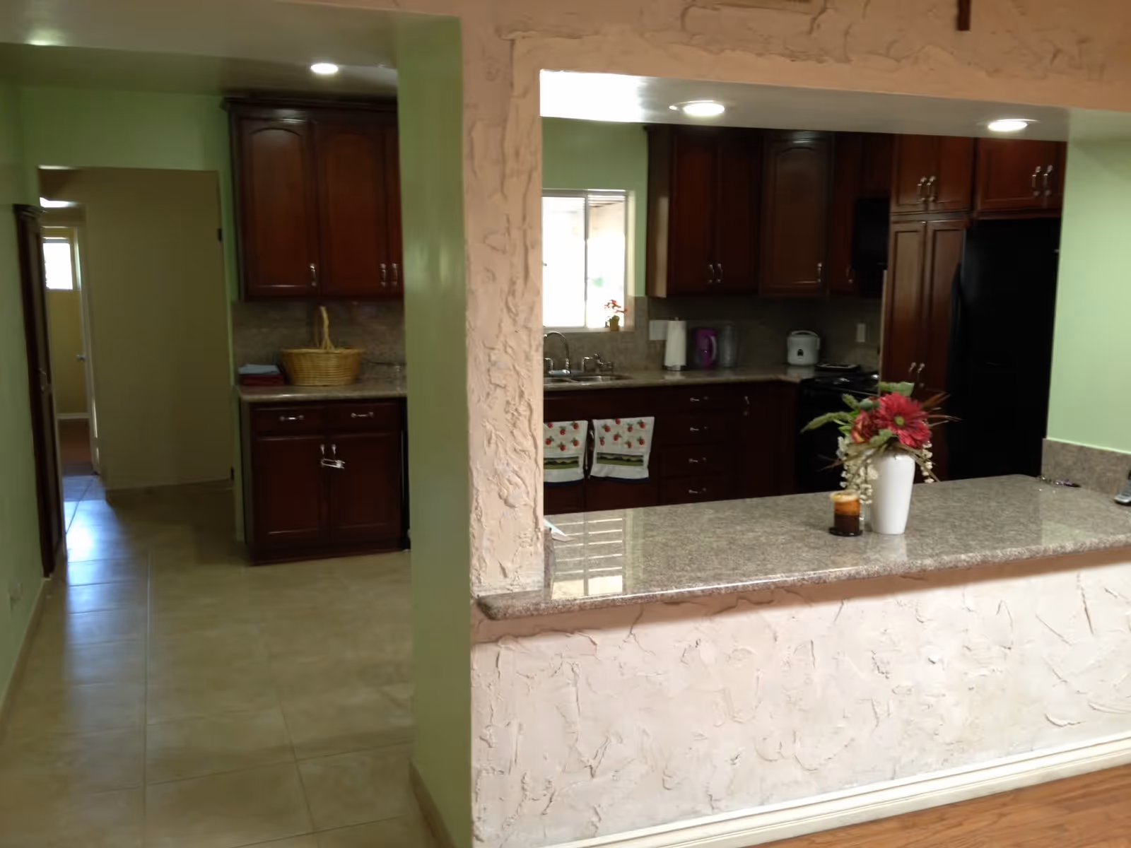 Interior view of a kitchen with dark wooden cabinets, a granite countertop with a vase of flowers, and kitchen appliances including a black refrigerator and a stove. The kitchen is visible through a pass-through window with textured walls, and there is a hallway to the left leading to other rooms.