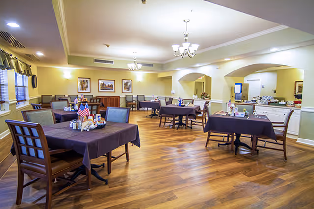 A spacious dining room with several tables covered in dark tablecloths, each set with condiments and small decorations. The room has wooden flooring, beige walls, and multiple chandeliers hanging from the ceiling. There are framed pictures on the walls and windows with valances on the left side.