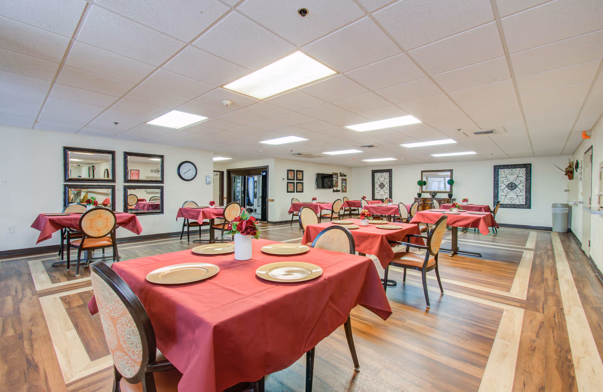 A spacious dining room with multiple tables covered in red tablecloths, each set with plates and a small floral centerpiece. The room has wood-patterned flooring, white walls decorated with framed mirrors and artwork, and bright overhead lighting.