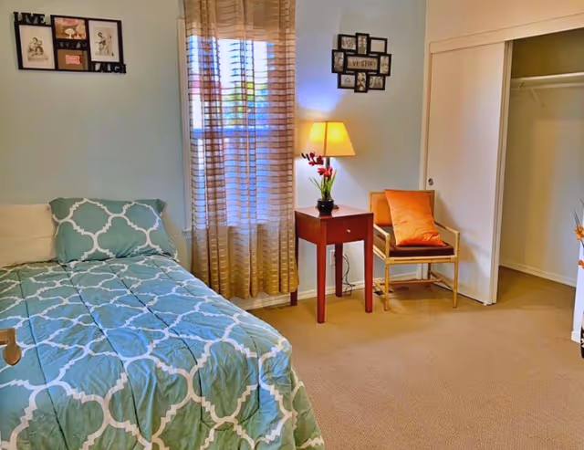 A cozy bedroom with a single bed covered in a blue and white patterned comforter. Next to the bed is a wooden nightstand with a lamp and a small vase of flowers. A wooden chair with an orange cushion is placed near a closet with sliding doors. The room has beige carpet and light-colored walls with framed pictures hanging. A window with sheer curtains allows natural light to enter the room.