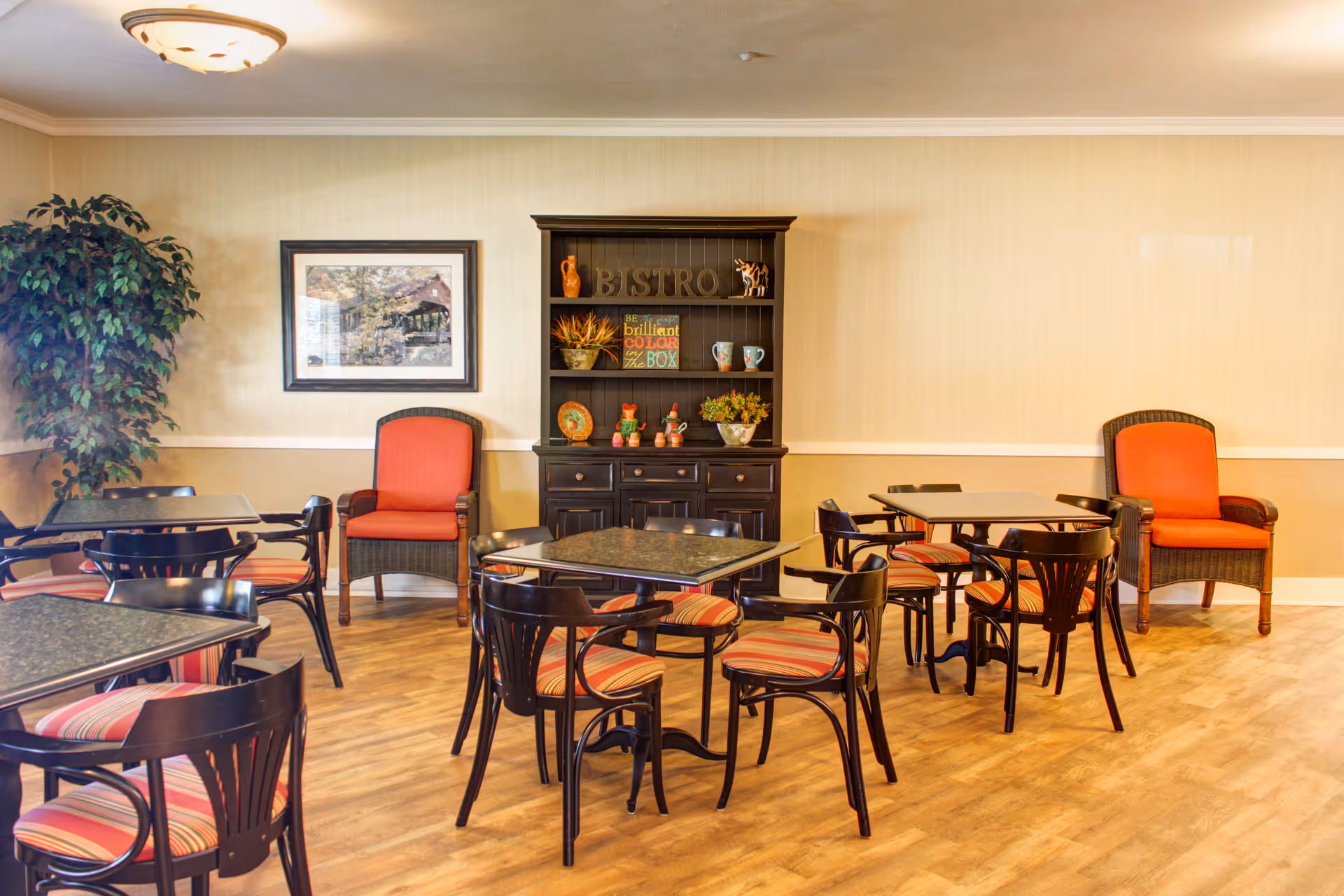 A dining area in a senior living facility with several square tables surrounded by black chairs with striped cushions. Two large armchairs with orange cushions are placed against the wall. A black hutch with decorative items and a sign that reads 'BISTRO' is centered on the wall. A framed picture and a large potted plant are also visible. The room has wooden flooring and beige walls.