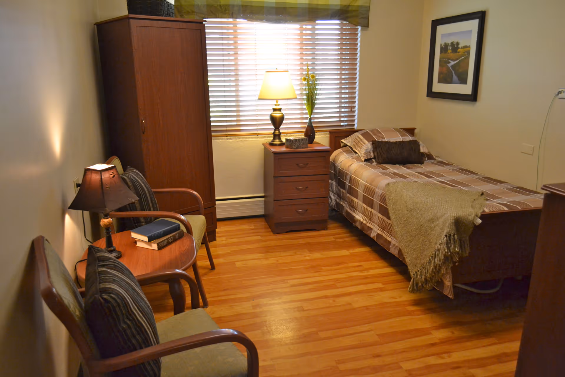 A cozy bedroom with a single bed covered in a plaid blanket and a brown throw. Next to the bed is a wooden nightstand with a lamp and a vase with flowers. There is a wooden wardrobe against the wall and two chairs with cushions around a small round table with two books and a lamp. The room has wooden flooring and a window with blinds and a green valance. A framed picture hangs on the wall above the bed.