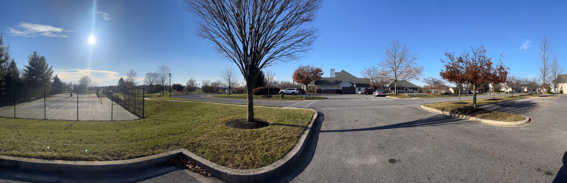 Panoramic view of an outdoor area at Carroll Vista featuring a fenced pickleball court on the left, a parking lot with several cars, leafless trees, and a building in the background under a clear blue sky with the sun shining brightly.