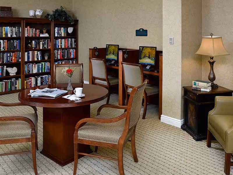 A cozy room with a round wooden table surrounded by four upholstered chairs. On the table, there is a vase with a red flower, a cup and saucer, and some papers. In the background, there are bookshelves filled with books and a computer workstation with two computers and chairs. To the right, there is a small side table with a lamp and a few stacked books, next to a green armchair.