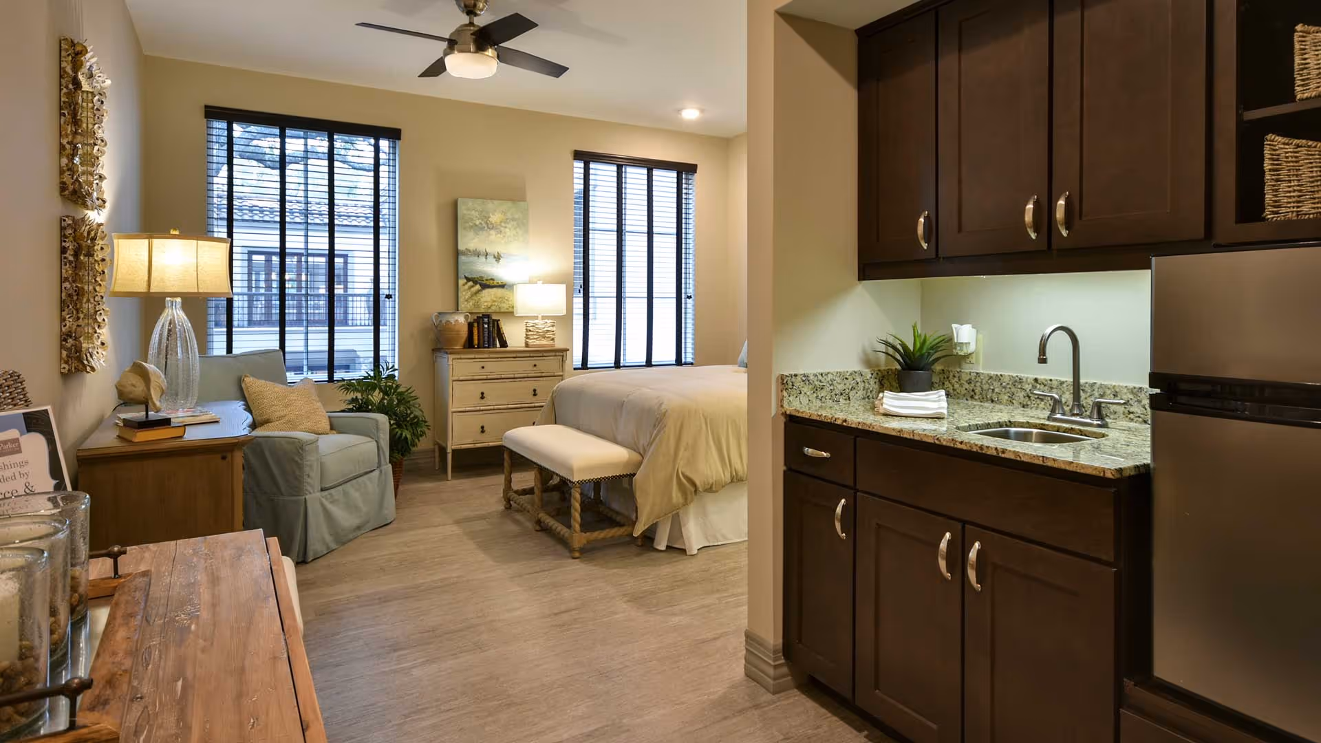 Interior view of a senior living facility room featuring a small kitchenette with dark wood cabinets, granite countertop, and a stainless steel mini refrigerator on the right. The room opens into a cozy bedroom area with a bed, a cushioned bench at the foot of the bed, a light blue armchair, a wooden dresser, and two large windows with blinds letting in natural light. The room is warmly lit with a ceiling fan light and a table lamp.