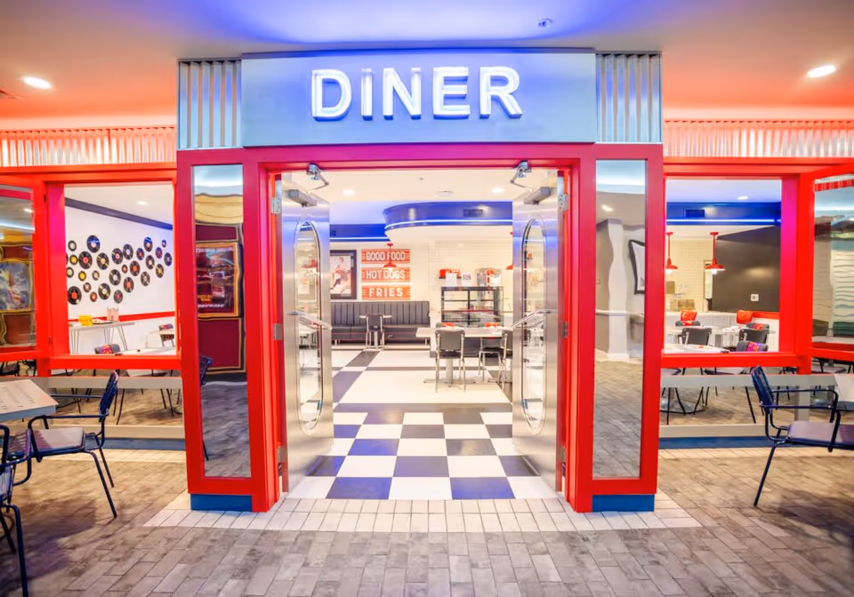 Entrance to a retro-style diner with red framing and double glass doors. Inside, there is a black and white checkered floor, tables and chairs, and a wall decorated with vintage signs and records. The word DINER is prominently displayed above the entrance in illuminated letters.
