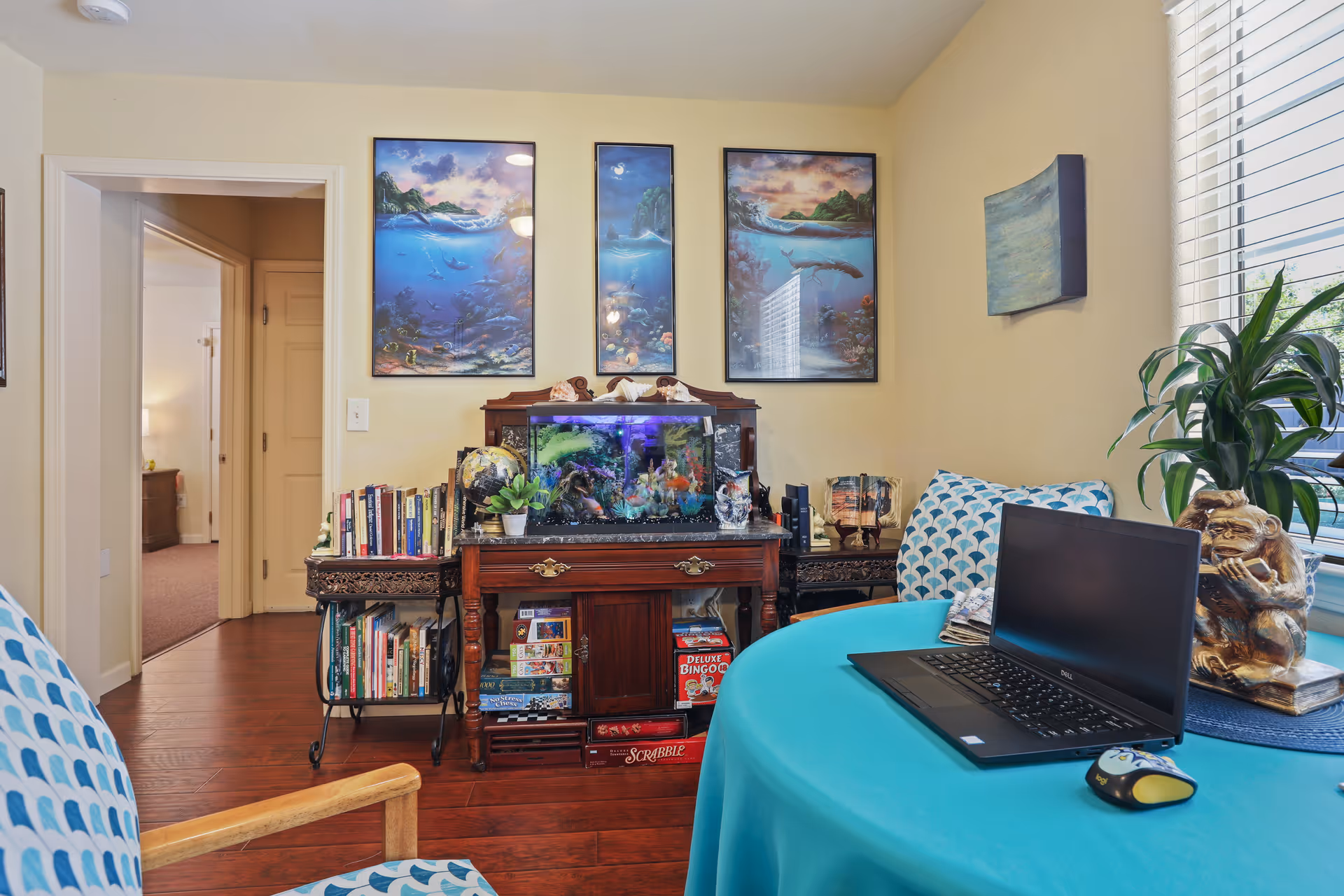 A cozy room with a round table covered by a turquoise tablecloth, a laptop and a computer mouse on top. Two chairs with blue and white patterned cushions are visible. Against the wall is a wooden cabinet with an aquarium, books, board games, and decorative items. Above the cabinet are three framed ocean-themed paintings. A window with blinds and a potted plant is on the right side, and an open doorway leads to another room.