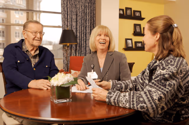 Three people sitting around a wooden table in a warmly lit room. An elderly man in a blue jacket, a woman with blonde hair wearing a gray blazer with a name badge, and another woman with brown hair in a patterned sweater are engaged in a cheerful conversation. A small floral arrangement is on the table, and framed photos are visible on the yellow wall behind them.