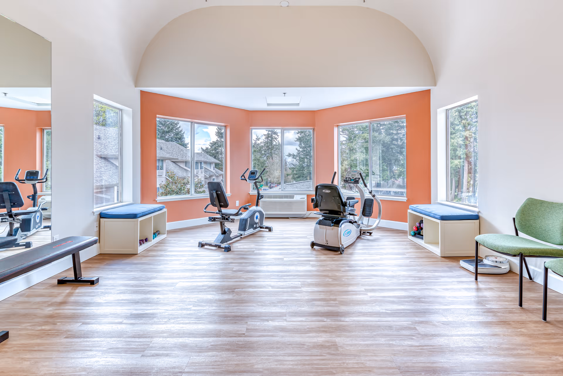 Bright exercise room with large windows showing outdoor trees and buildings. The room has light wood flooring, two recumbent exercise bikes, a bench, two cushioned window seats with storage underneath, and two green chairs. The walls are painted white with an orange accent wall around the windows.