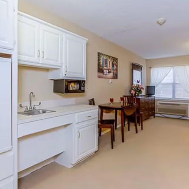 Studio-style senior living interior with a kitchenette on the left, a small dining table and chairs, and a dresser under a window.