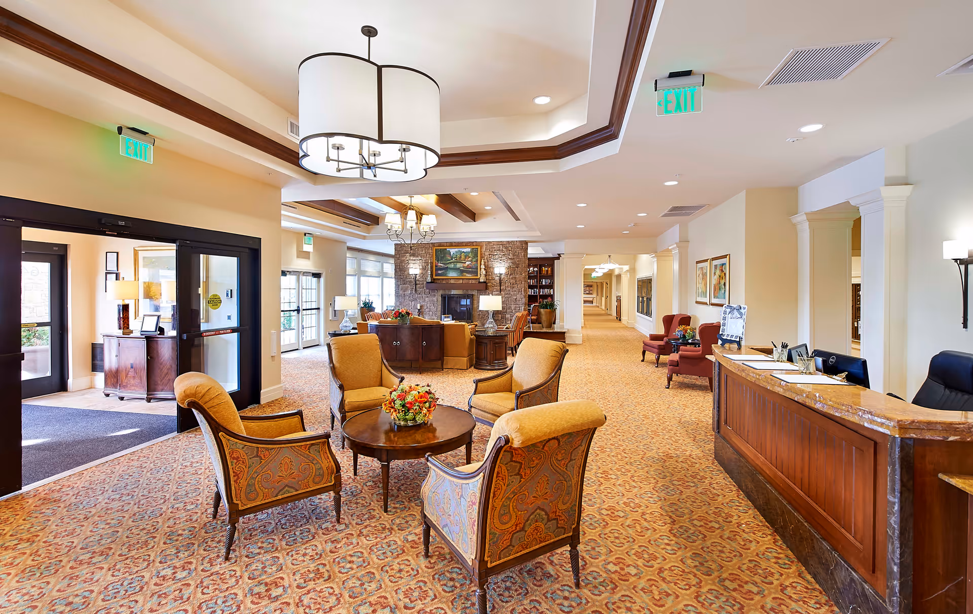 A spacious senior living facility lobby with patterned carpet, a seating area consisting of four upholstered armchairs around a round wooden table with a floral centerpiece, a wooden reception desk on the right, and a stone fireplace with a painting above it in the background. The area is well-lit with ceiling lights and chandeliers, and there are exit signs above the doors.