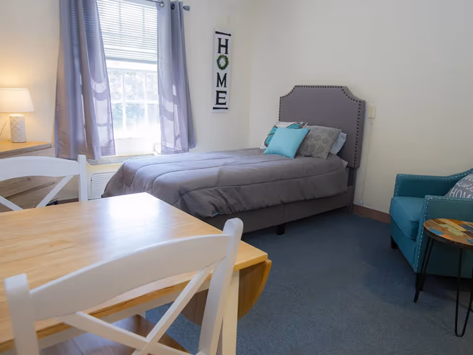 A bright studio bedroom with a single bed, small dining table, lamp and teal armchair beside a window.