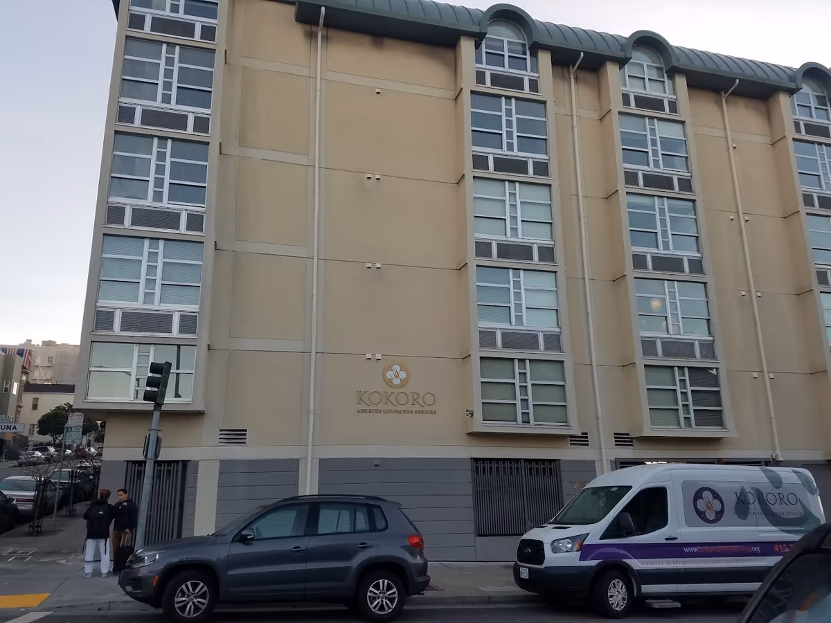 Exterior view of a multi-story beige building with large windows and a sign that reads 'Kokoro Assisted Living for Seniors'. Two vehicles are parked in front, including a van with the Kokoro logo and website. Two people are standing on the sidewalk near a traffic light.