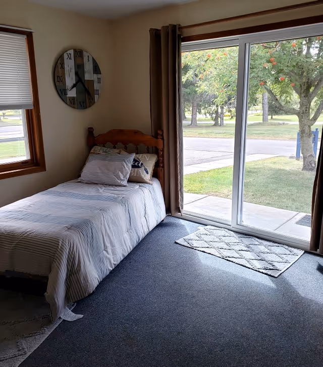 Small bedroom with a single bed, wall clock, and sliding glass door opening to a grassy outdoor view.