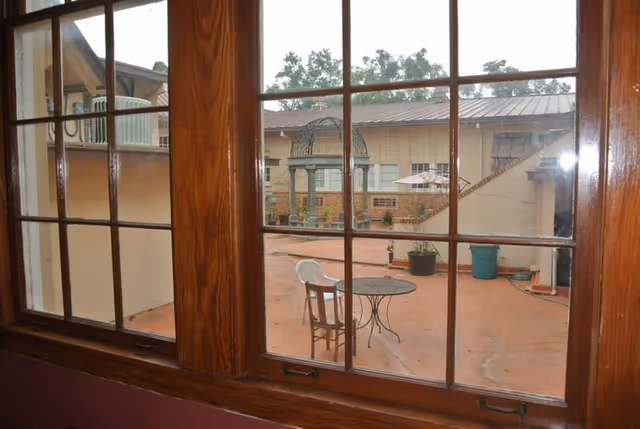 View through a wooden-framed window showing an outdoor patio area with a round metal table, a wooden chair, a white plastic chair, potted plants, and a decorative metal gazebo structure. The patio is surrounded by beige buildings with windows and a metal roof.