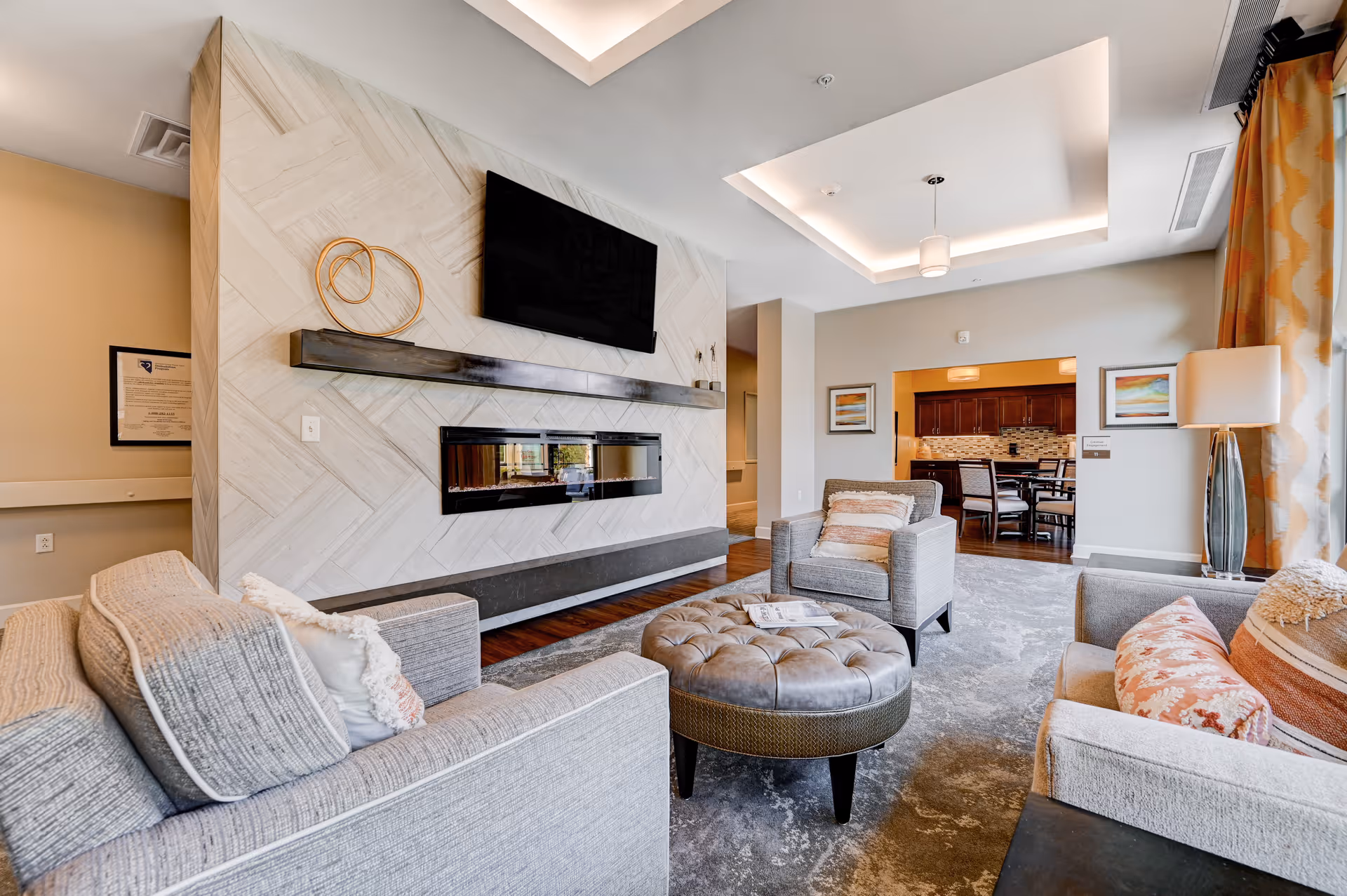 Bright, modern living room with sofas and armchairs arranged around a tufted ottoman, a wall-mounted TV above a linear fireplace, and a view into a dining area.