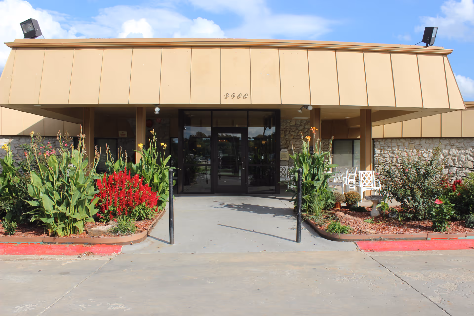 Front entrance of a building with beige paneling and stone walls, featuring glass double doors. There are flower beds with green plants and red flowers on either side of the entrance, and white chairs are visible on the right side under the covered area.