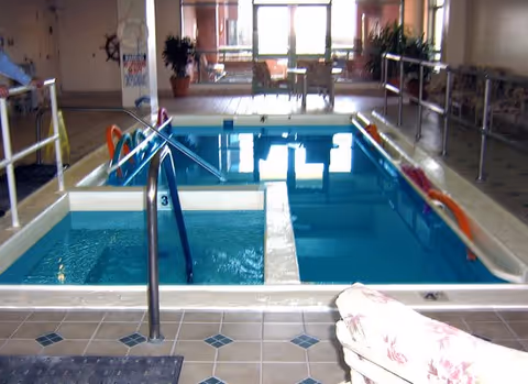 Indoor pool area with a small hot tub section and a larger swimming pool, surrounded by tiled flooring and metal handrails. There are chairs and plants in the background near large windows letting in natural light.