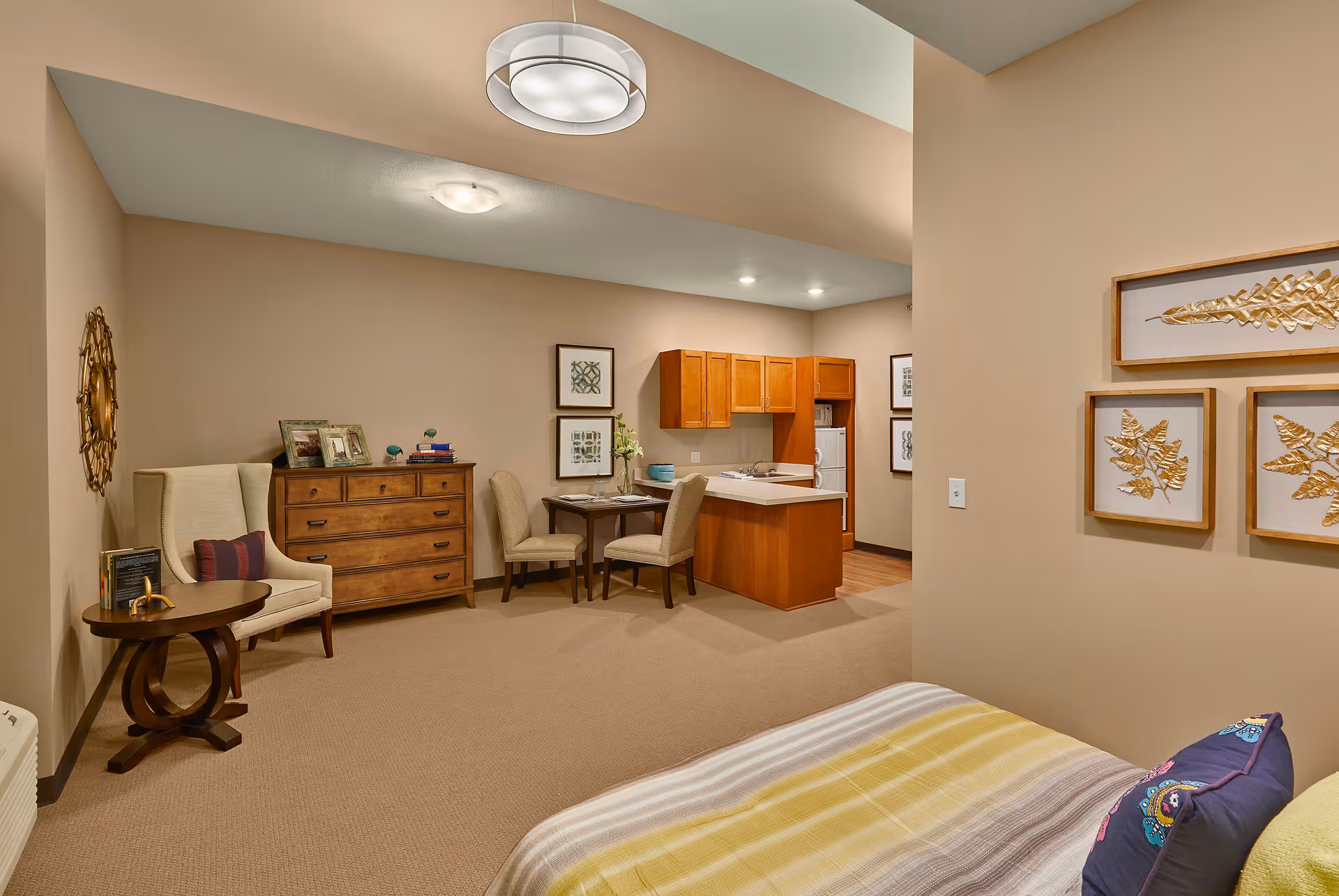 Interior view of a nursing home room featuring a bed with a striped yellow and gray blanket and a decorative pillow, a small wooden table with a beige armchair, a wooden dresser with framed photos and books, a small dining table with two chairs, and a kitchenette with wooden cabinets and a white refrigerator. The walls are beige with framed leaf artwork and decorative pieces.