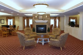 A cozy senior living common area featuring four patterned armchairs arranged around a round wooden coffee table in front of a stone fireplace. The room has red patterned carpet, large windows with curtains, and additional seating with wooden chairs and tables in the background.
