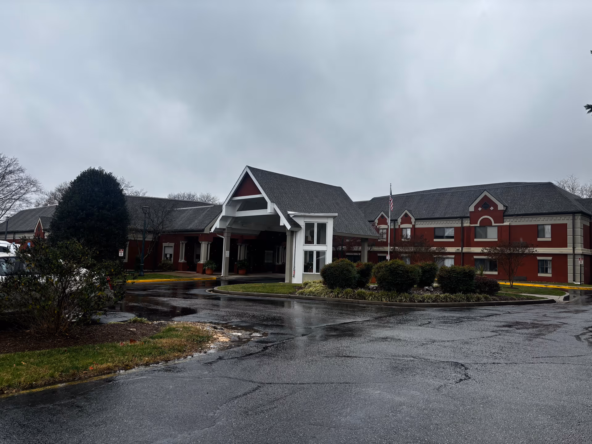 Exterior view of a senior living community building with a covered entrance, red walls, and a gray roof on a cloudy day with wet pavement and some landscaping including bushes and trees.