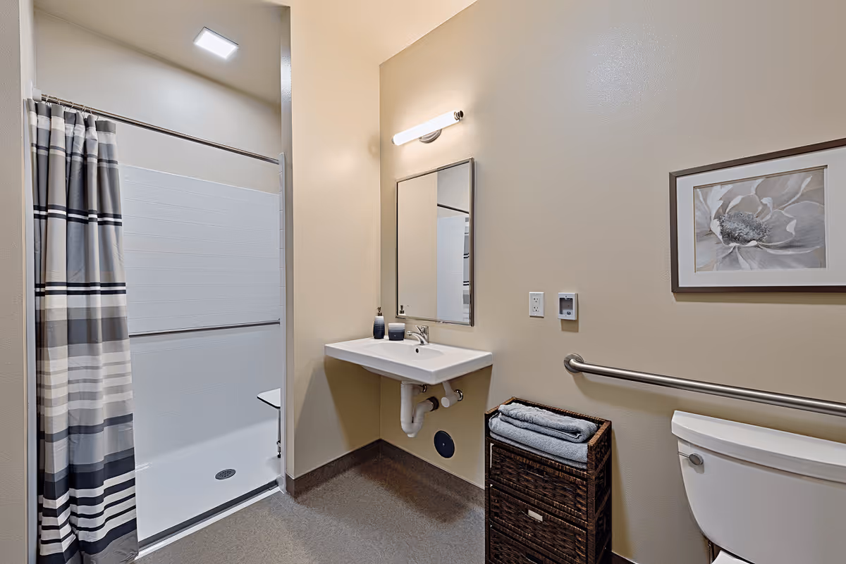 A clean and accessible bathroom featuring a walk-in shower with a striped shower curtain, a wall-mounted sink with soap dispensers, a mirror with a light above it, a toilet with a grab bar, a wicker storage basket with folded towels, and a framed flower artwork on the wall.