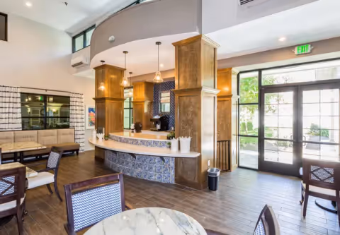 Interior view of a bright and spacious common area in The Windham facility featuring a curved counter with decorative tiles, wooden pillars, pendant lights, seating with chairs and a bench, large windows and glass doors allowing natural light to enter, and a view of greenery outside.