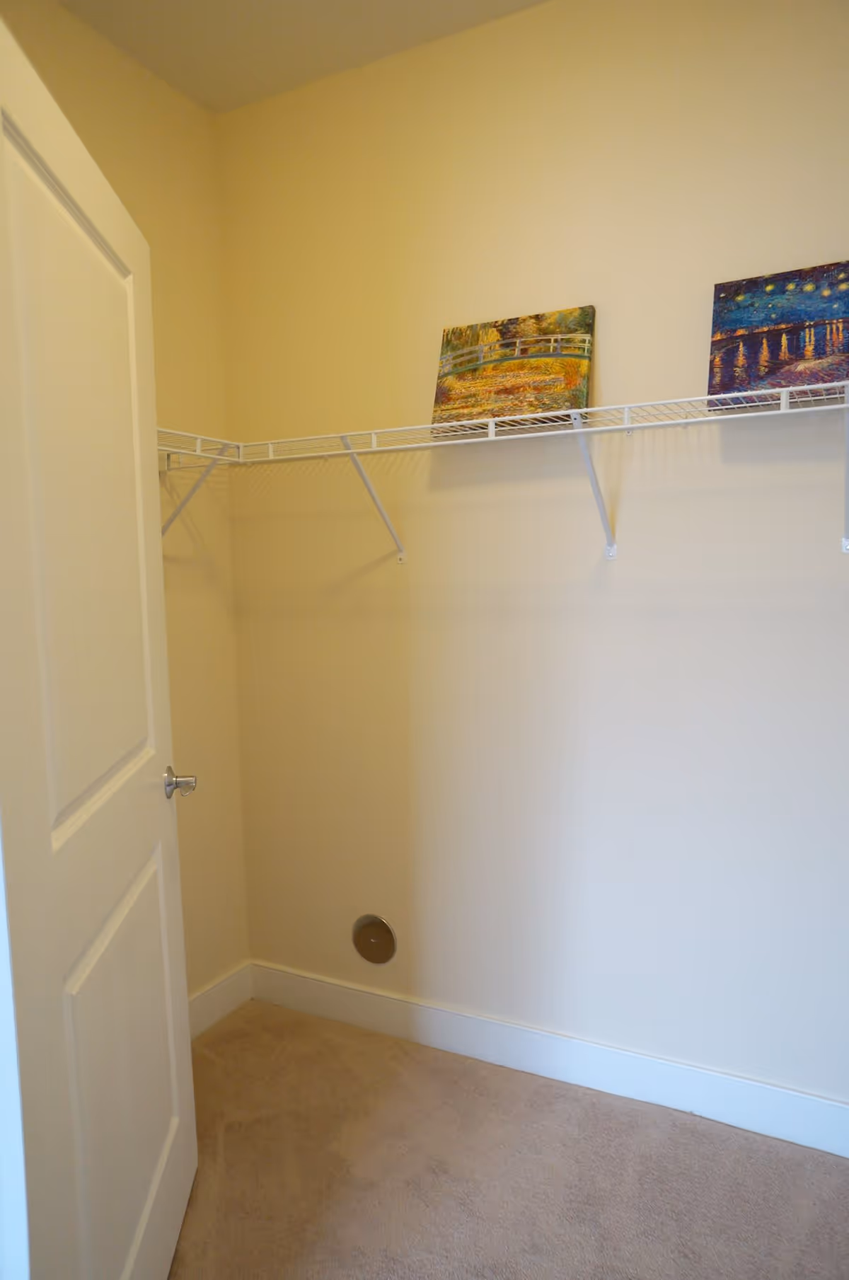 Empty walk-in closet with beige walls and carpeted floor. A white wire shelf runs along two walls near the ceiling, holding two small paintings. A white door with a silver handle is partially open.